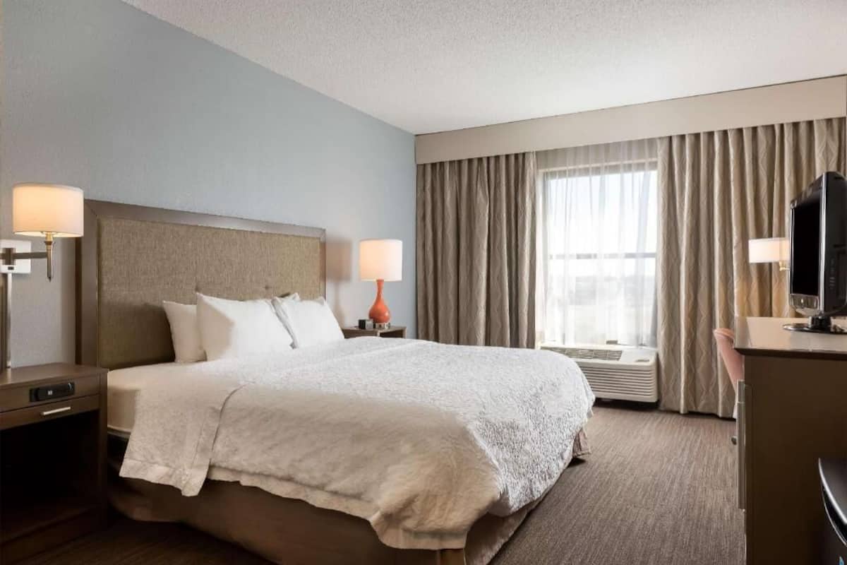 A hotel room with a large bed, two bedside tables with lamps, a TV, and a window with curtains.