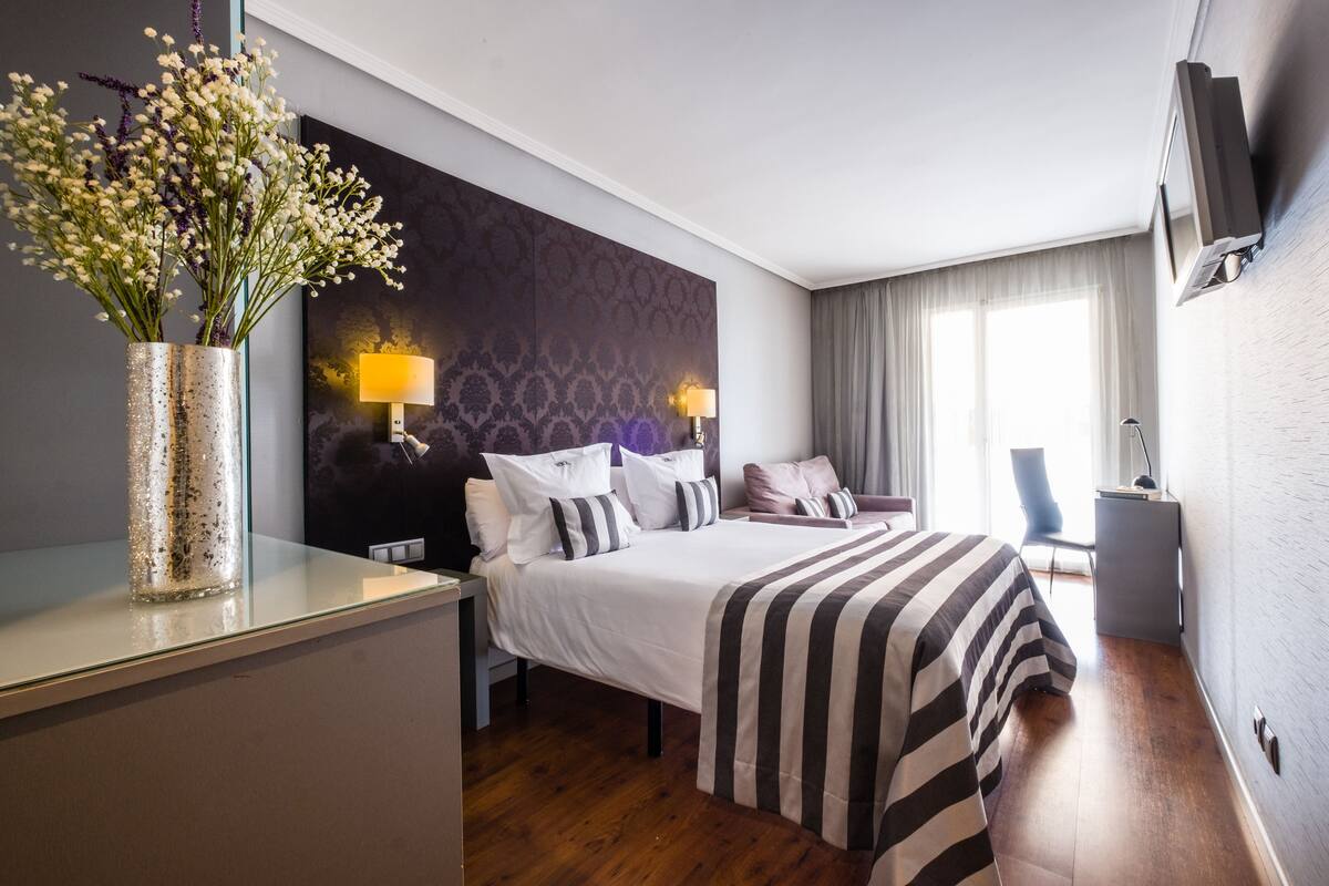 A hotel room with a bed, bedside lamps, a TV, a desk with a lamp, and a vase with flowers.