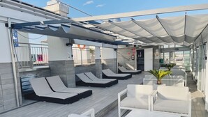A rooftop lounge area with sun loungers and white furniture, featuring a covered patio and a view of the city.