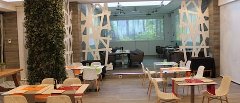 A modern dining area with multiple tables and chairs, a large decorative plant, and a projection screen in the background.