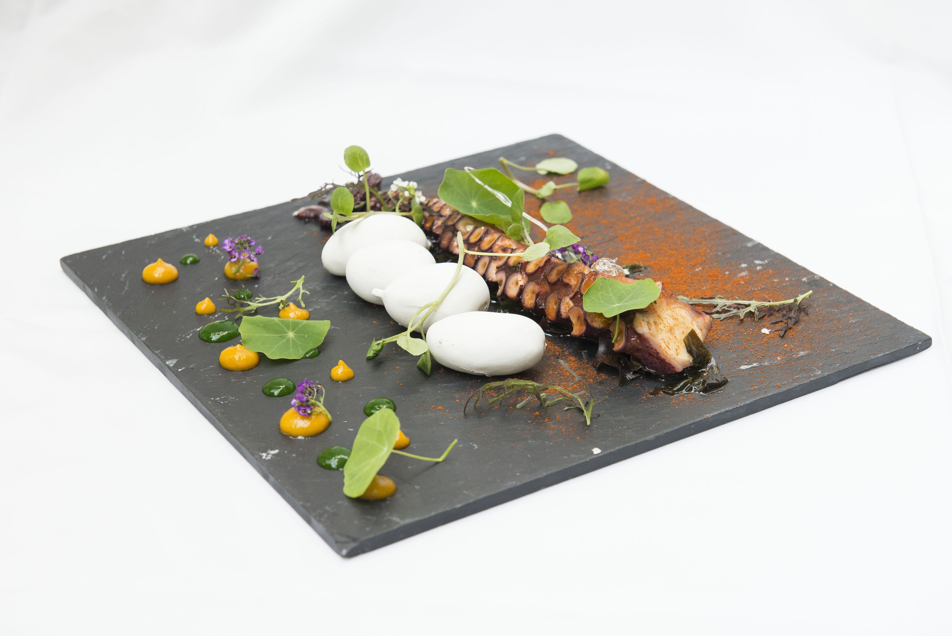 Una tabla de pizarra cuadrada con un plato que presenta pescado, esferas blancas y hojas verdes.