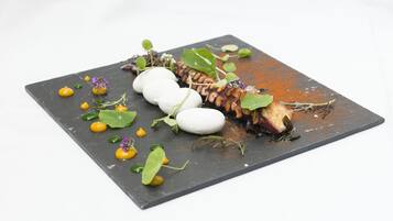 Una tabla de pizarra cuadrada con un plato que presenta pescado, esferas blancas y hojas verdes.