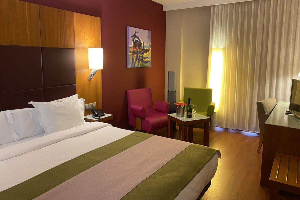 A hotel room with a large bed, a desk with a chair, a small table with a bottle and glasses, a phone, and a painting on the wall.