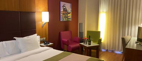 Quarto de hotel com uma cama grande, uma escrivaninha com cadeira, uma mesinha com garrafa e copos, um telefone e um quadro na parede.