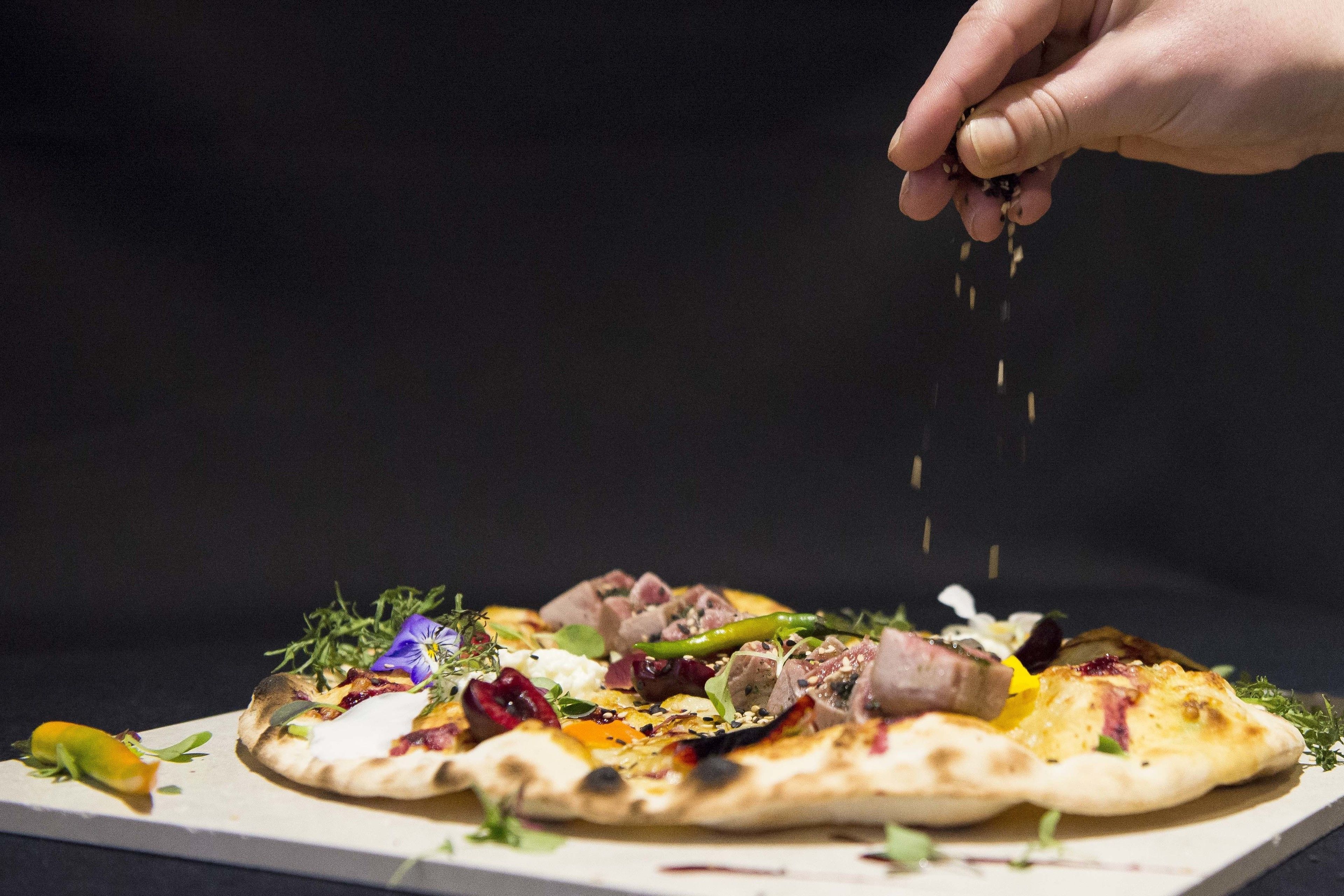 A pizza with various toppings and a hand sprinkling seasoning on it.