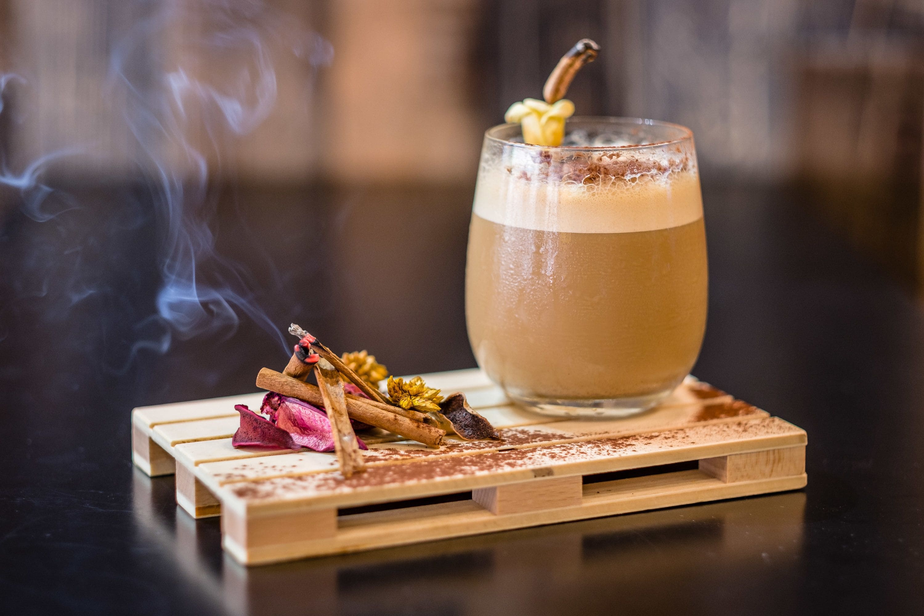 Un vaso de una bebida marrón claro, adornado con una rama de canela, sobre un posavasos de madera con flores secas y palitos de canela.