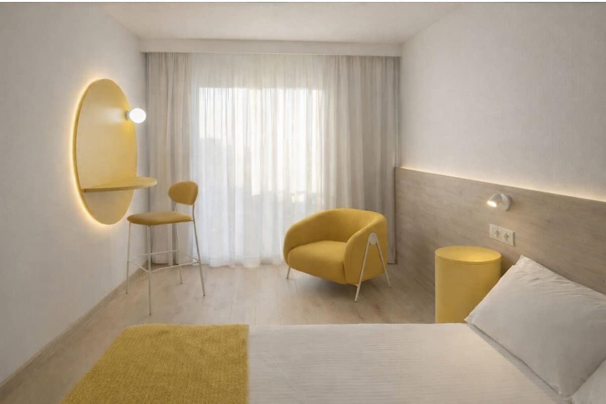 A hotel room with a bed, a yellow armchair, a small table, a mirror, and a window with sheer curtains.