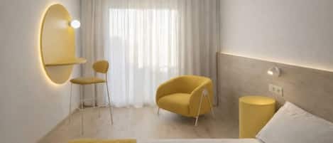 A hotel room with a bed, a yellow armchair, a small table, a mirror, and a window with sheer curtains.