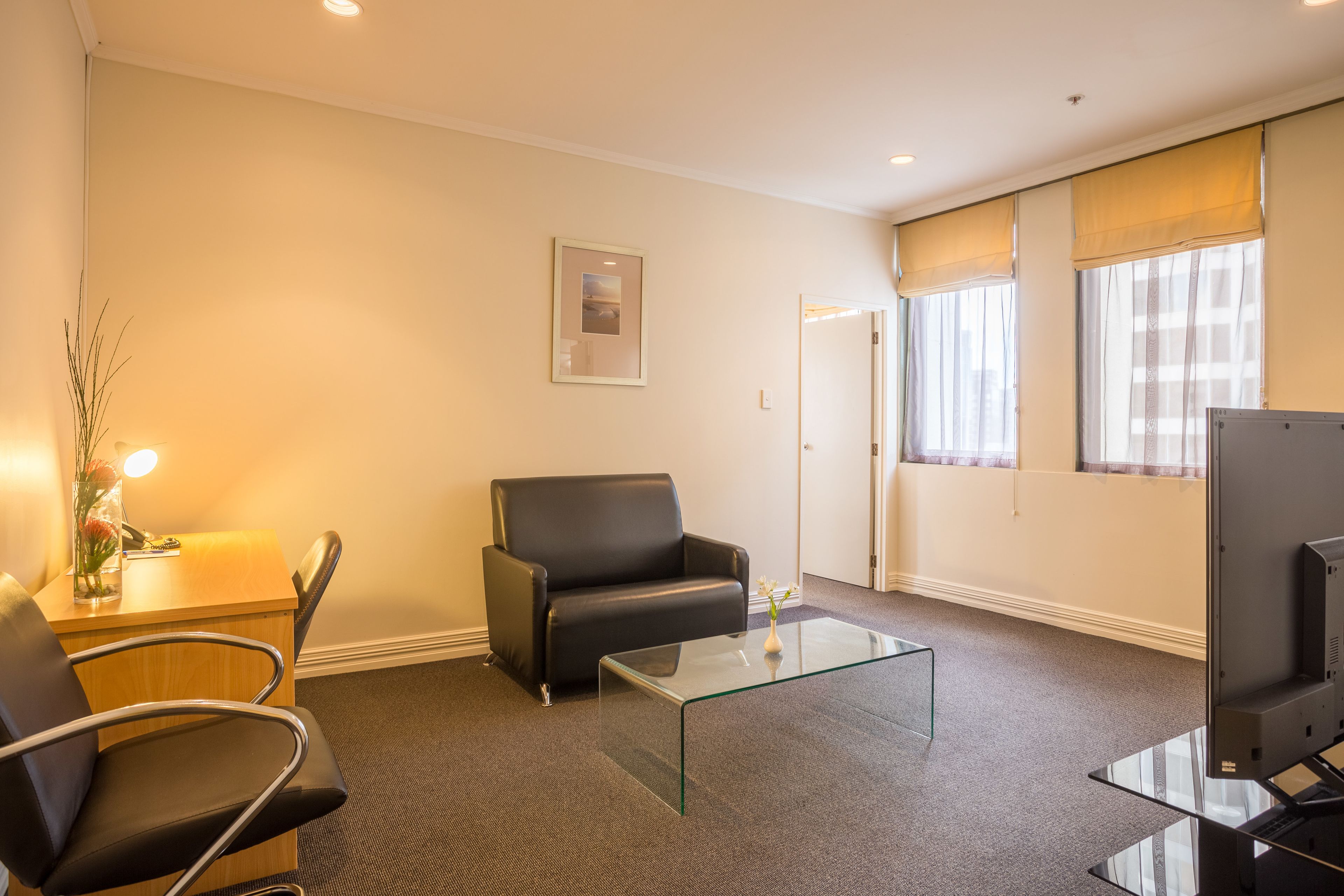 Une chambre d’hôtel avec une table basse en verre, deux chaises noires, un bureau avec une lampe, un tableau encadré au mur et un téléviseur.