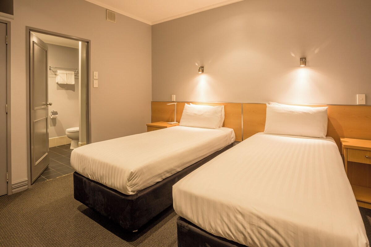 Habitación de hotel con dos camas individuales, cabecera de madera, mesitas de noche, baño con inodoro y perchero, y puerta que da al pasillo.
