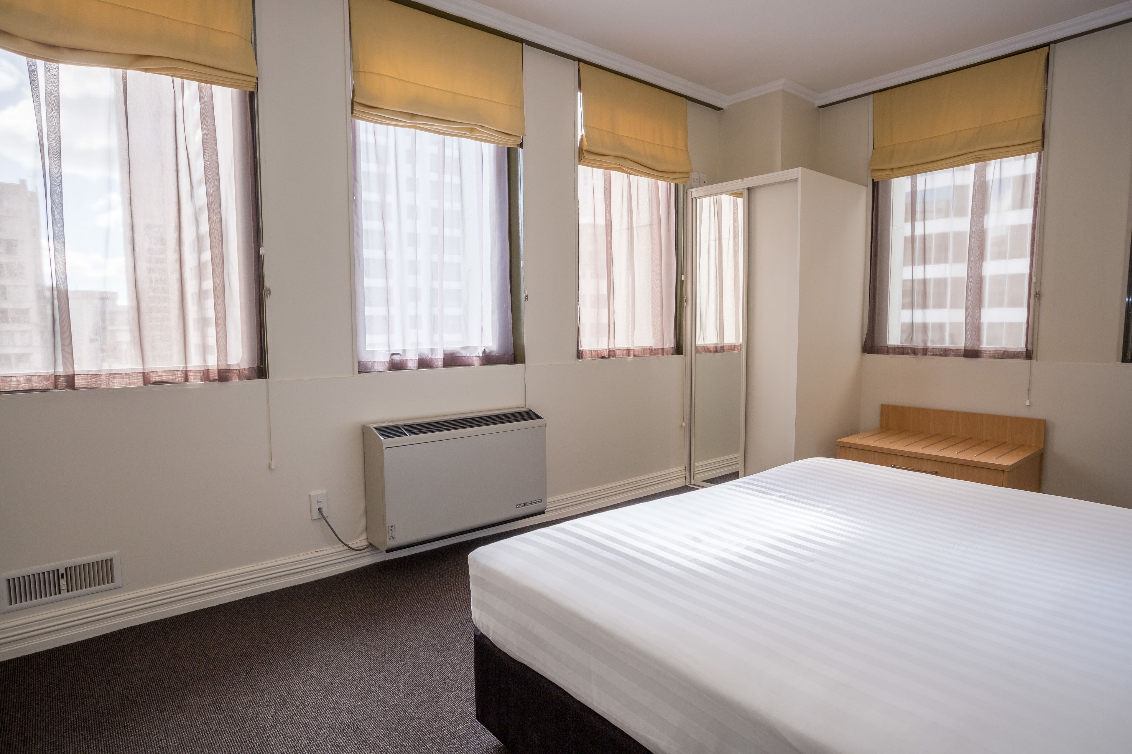 Hotellihuone, jossa on suuri sänky, puinen penkki, patteri ja ikkunoissa verhot.
