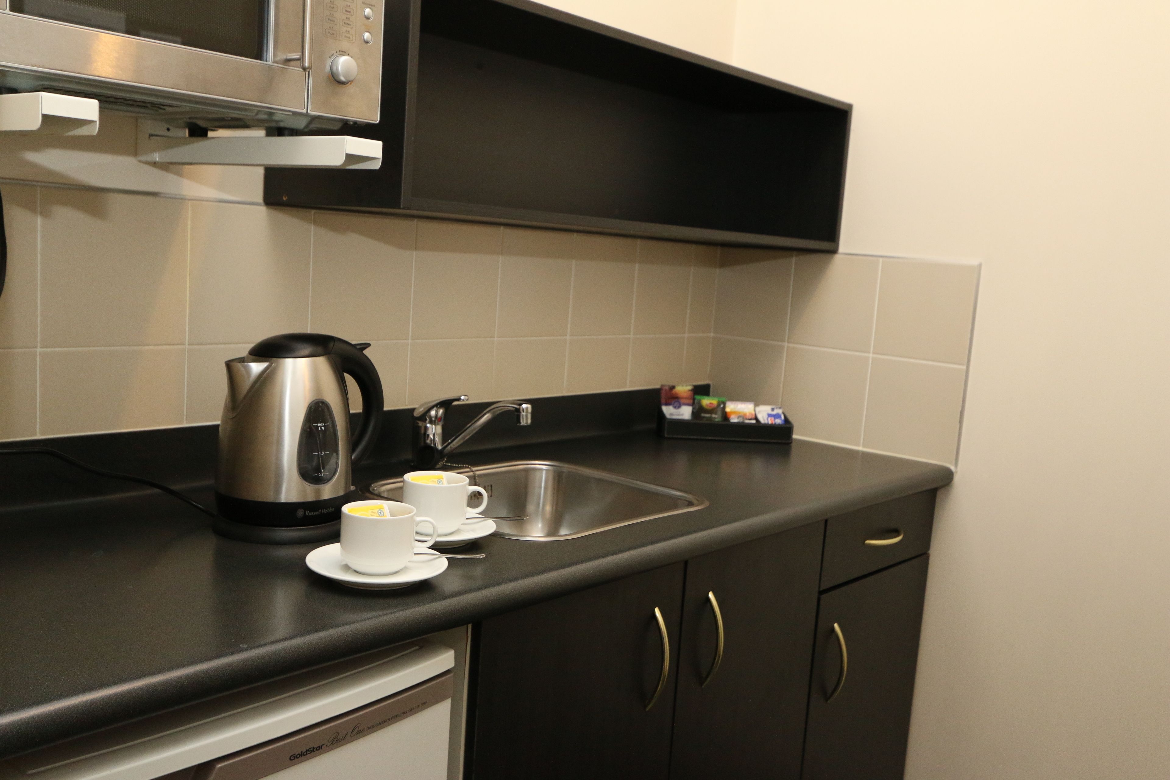 A modern kitchen with a stainless steel kettle, two white cups, a sink, and a microwave above the counter.