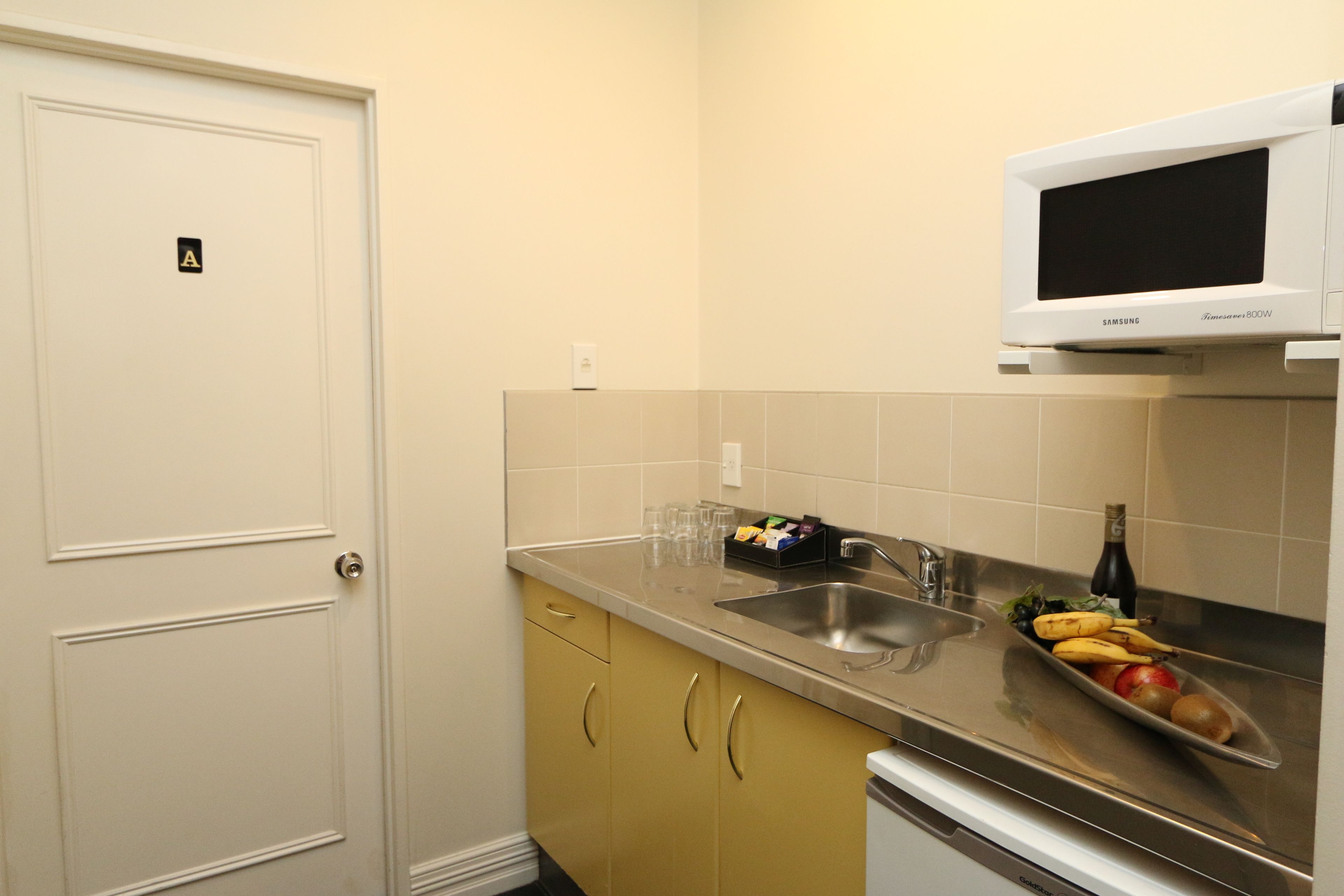 A compact kitchen with a microwave, sink, and a small countertop with fruits and glasses.