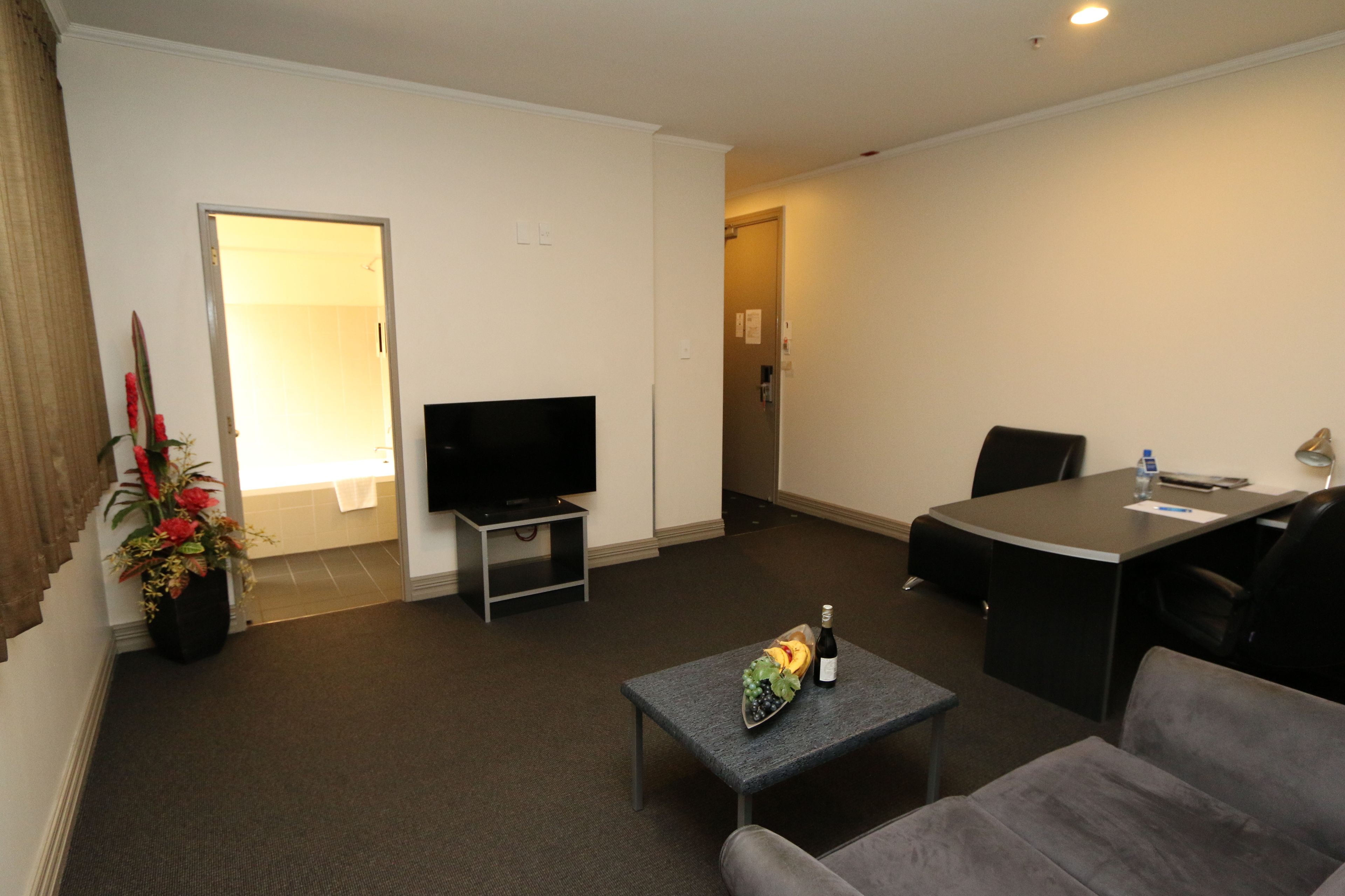Ein Hotelzimmer mit Flachbildfernseher, einem kleinen Tisch mit Flasche und Obstkorb, einer Couch, einem Esstisch mit Stühlen und einer Topfpflanze.