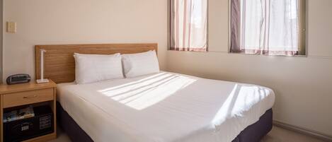 A neatly made bed in a hotel room with a wooden headboard, a bedside table with a lamp, and a window with sheer curtains.