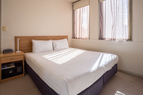 Una cama bien tendida en una habitación de hotel, con un cabecero de madera, una mesita de noche con lámpara y una ventana con cortinas translúcidas.