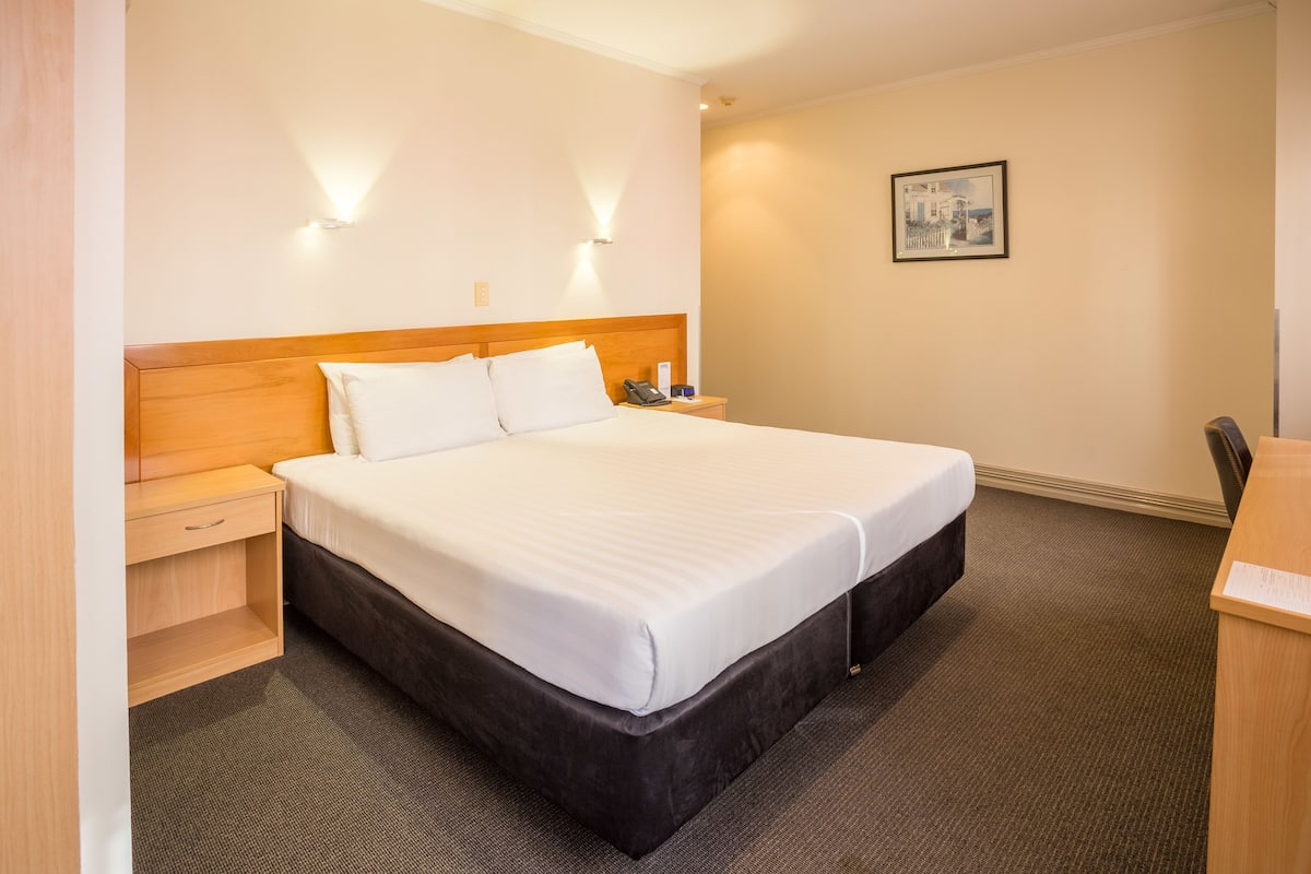 A hotel room with a large bed, a wooden nightstand, a desk, and a framed picture on the wall.