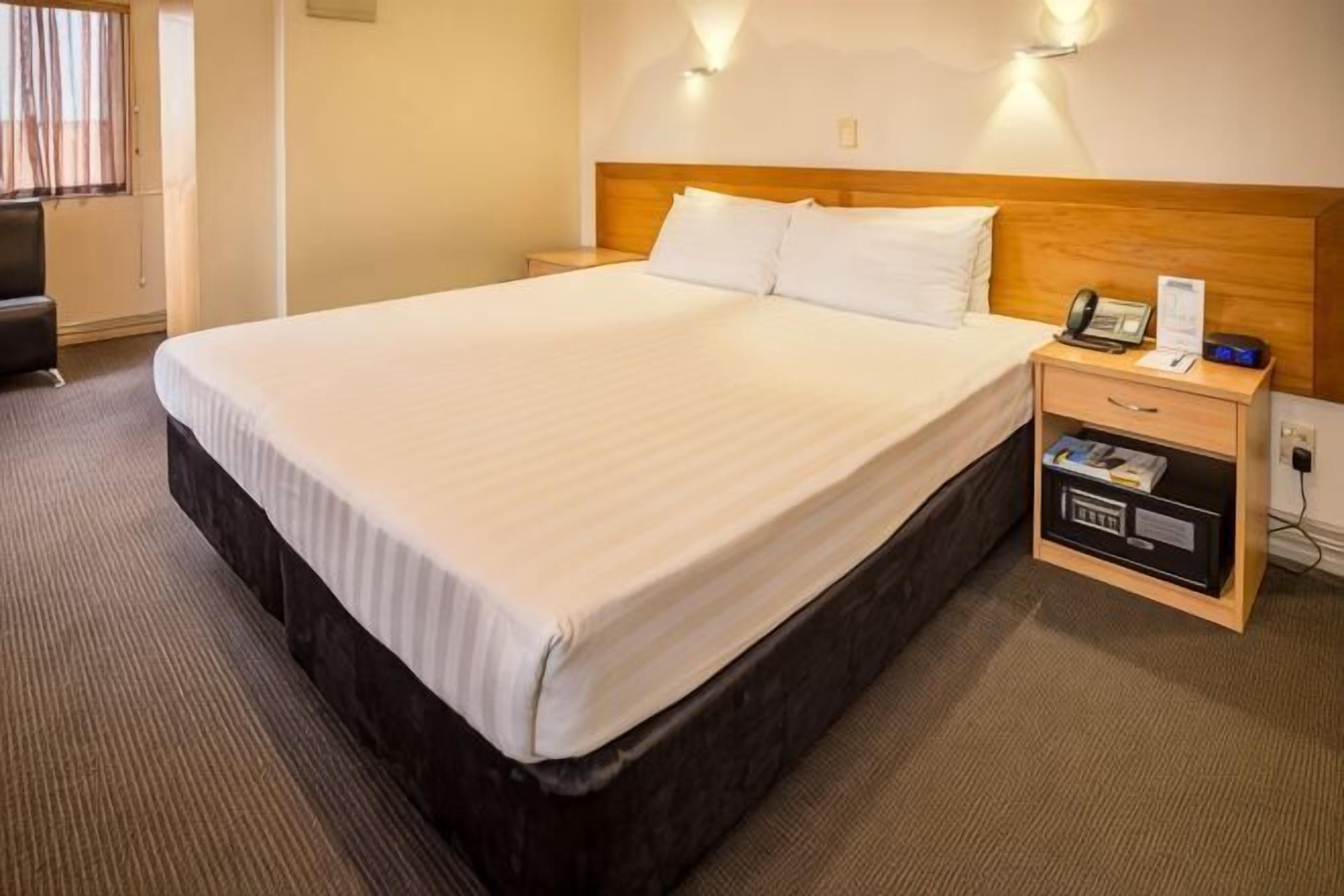 A hotel room with a large bed, a wooden headboard, a bedside table with a phone and a small shelf, and a wall-mounted light.