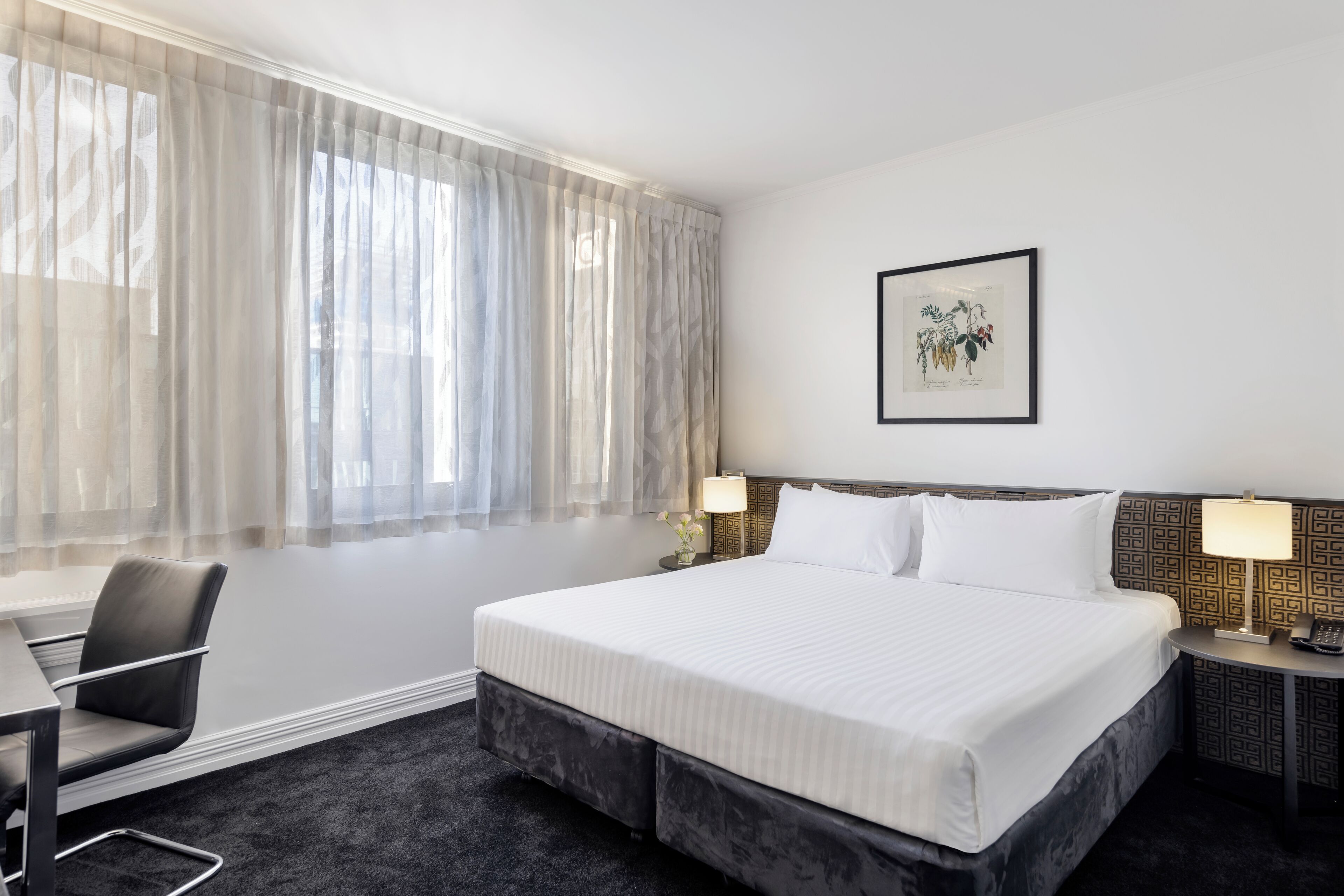 A hotel room with a large bed, a desk, and a chair. There is a window with sheer curtains and a framed botanical print on the wall.
