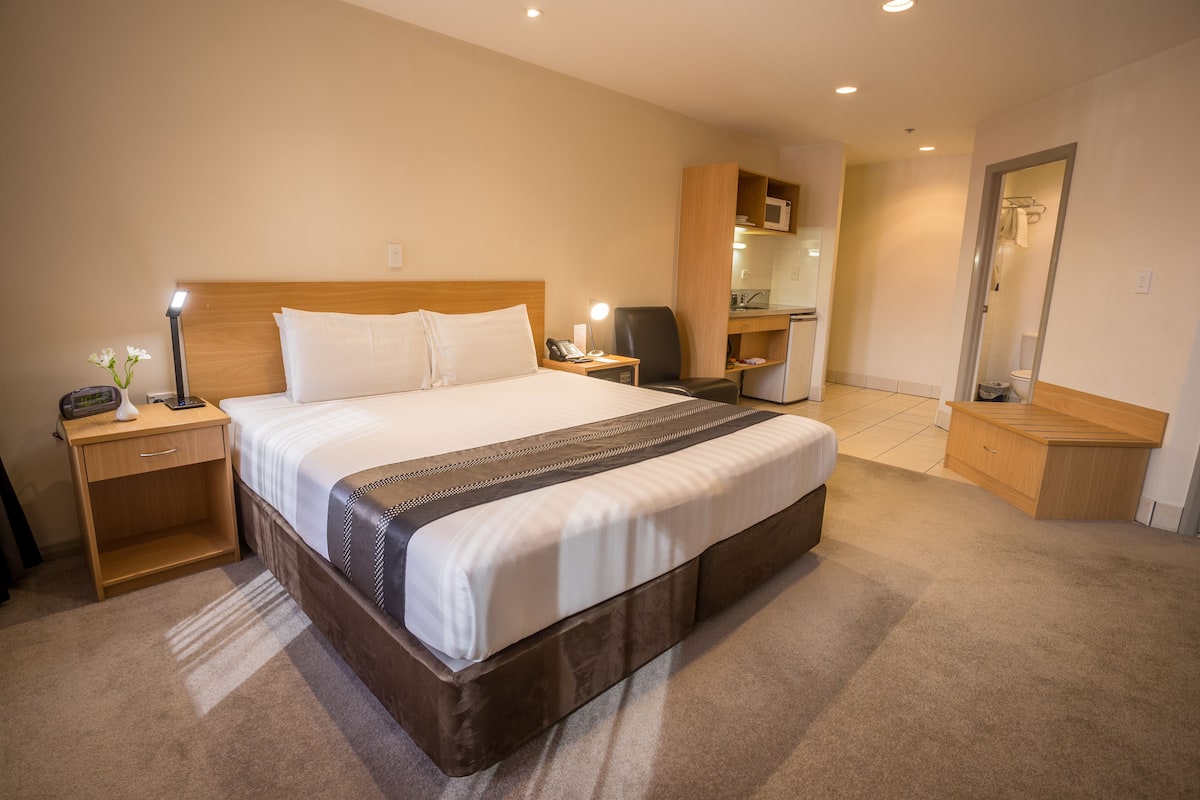 A hotel room with a large bed, bedside tables, a desk, a chair, a microwave, and a bathroom visible through an open door.