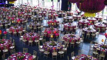 Een ruime balzaal met ronde tafels gedekt voor evenementen, met gouden stoelen en rode tafelkleden.