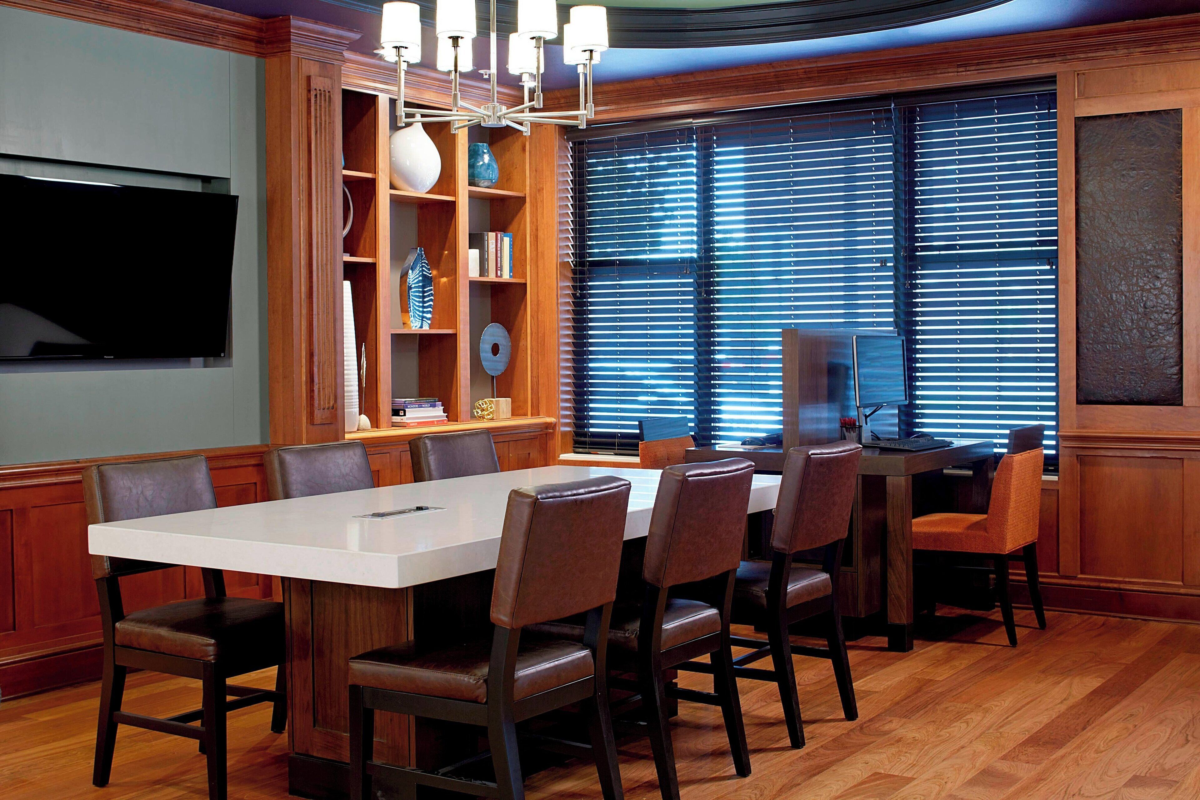 Un comedor con una gran mesa rectangular rodeada de sillas, una araña y una estantería con objetos decorativos.