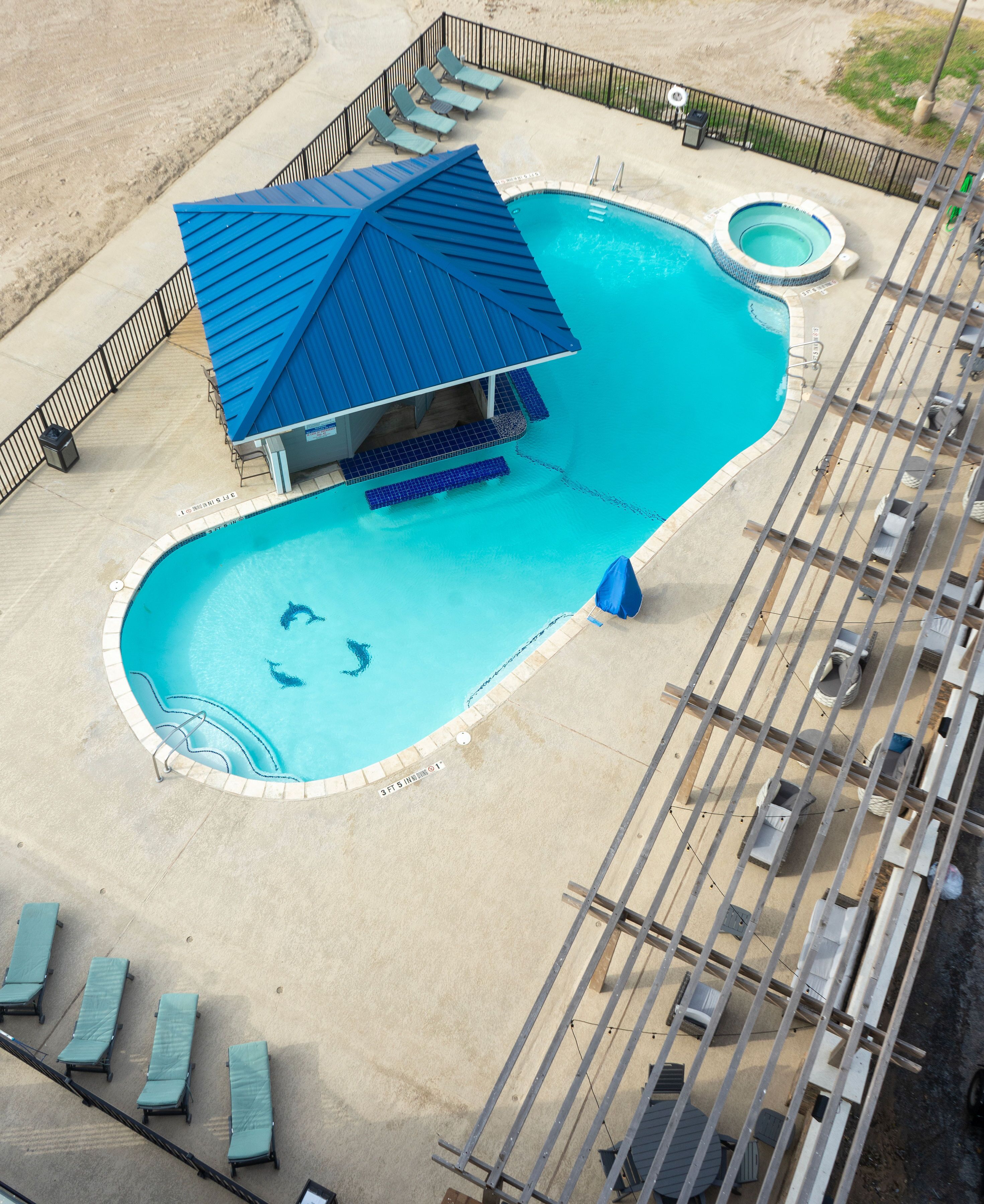 An aerial view of a swimming pool with a blue-roofed structure, lounge chairs, and a small hot tub.