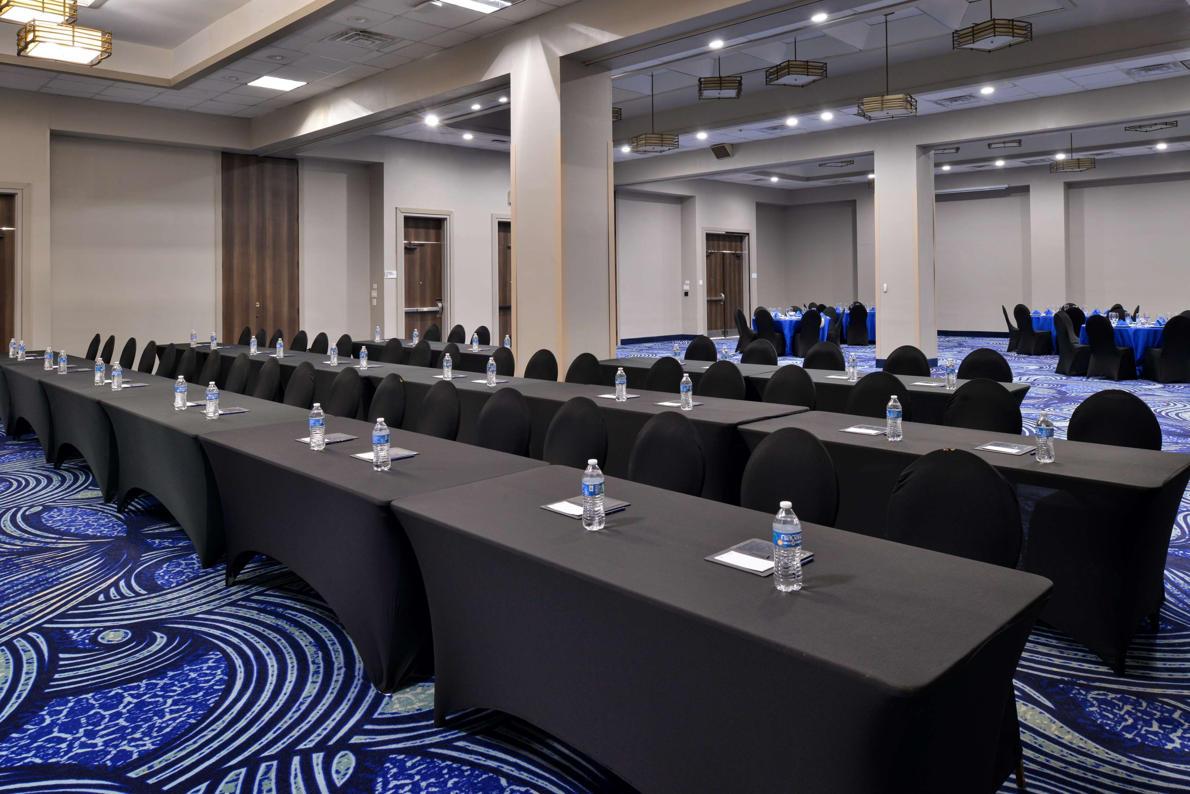 A conference room with long tables, chairs, water bottles, and a blue carpet.