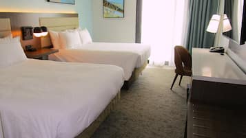Habitación de hotel con dos camas, un escritorio, una silla, una lámpara y un ventanal grande con cortinas.