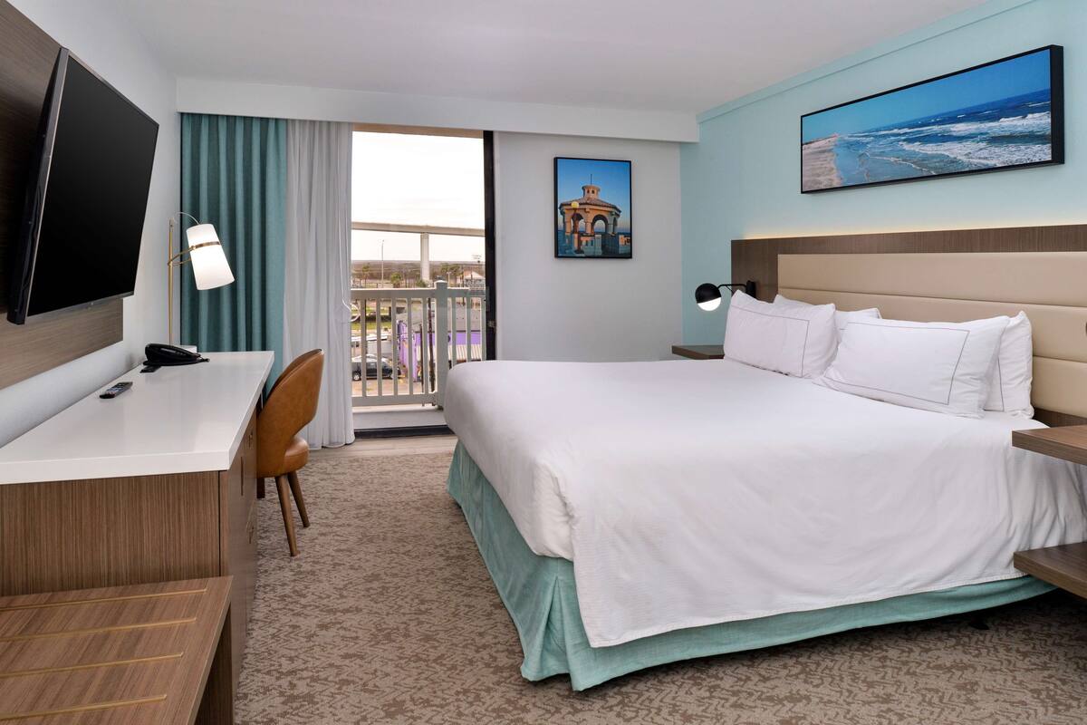 A hotel room with a bed, a desk, a chair, a TV, and a balcony with a view of the sea.