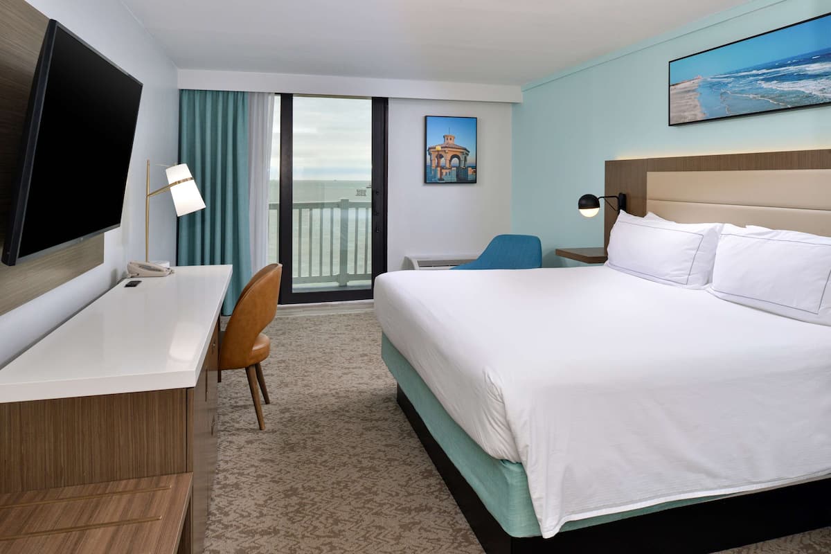 A hotel room with a bed, a desk, a chair, a TV, and a balcony with a view of the ocean.
