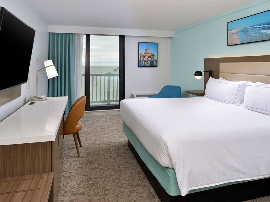 A hotel room with a bed, a desk, a chair, a TV, and a balcony with a view of the ocean.