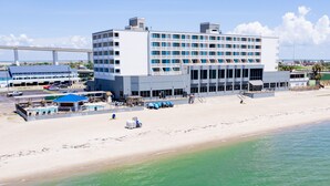 A large hotel with multiple floors and balconies, situated on a beach with a parking area and a pool.