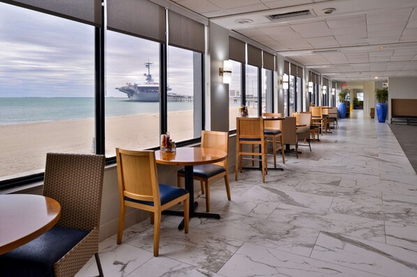 A restaurant with large windows offering a view of the beach and a pier. Tables and chairs are arranged along the walls.