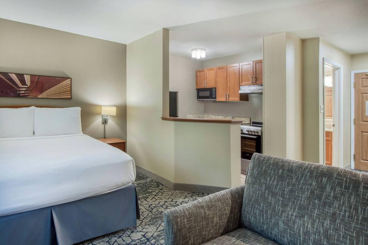 A hotel room with a bed, a seating area, a kitchenette, and a door leading to another room.