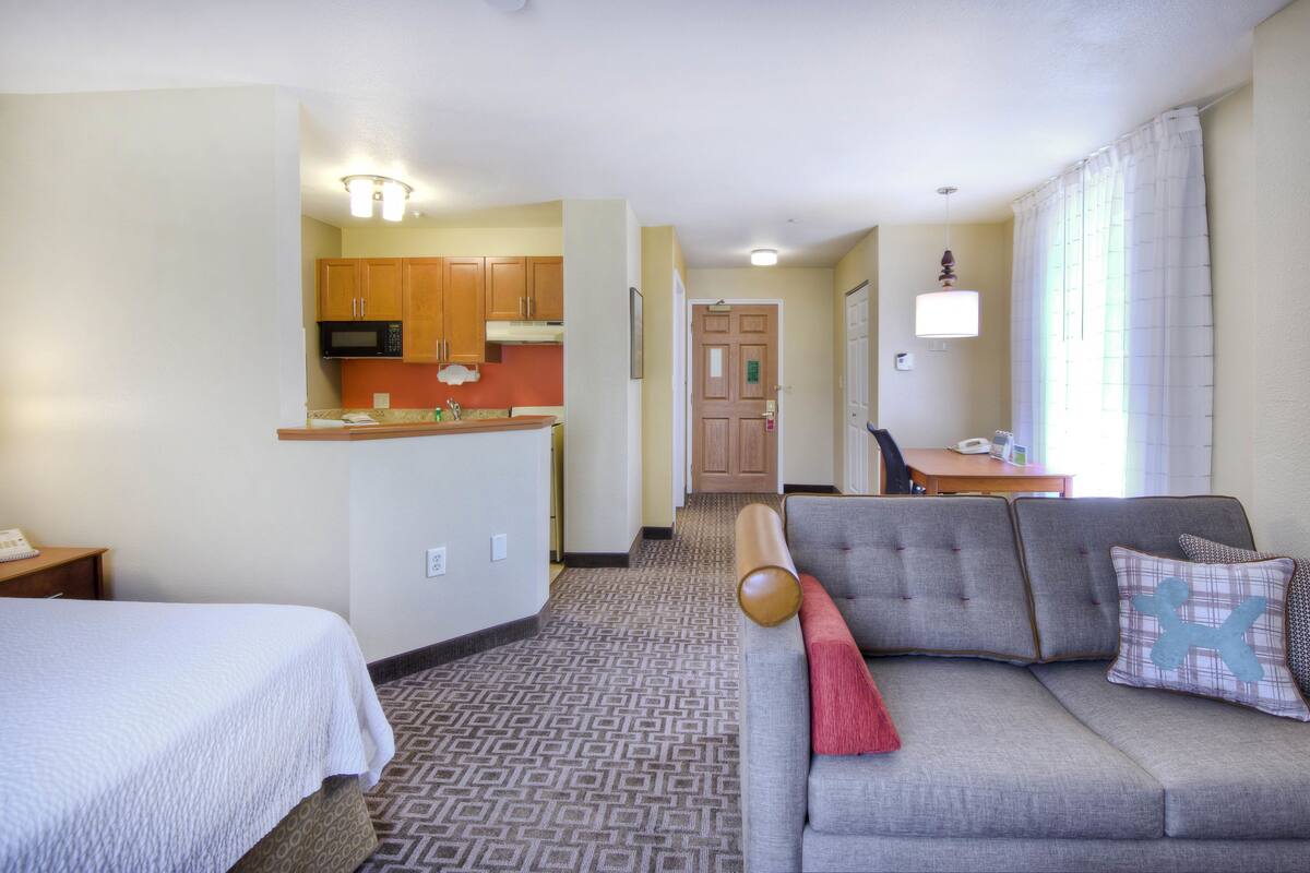 A hotel room with a kitchenette, a sofa with a red throw pillow, and a bed with a white comforter.
