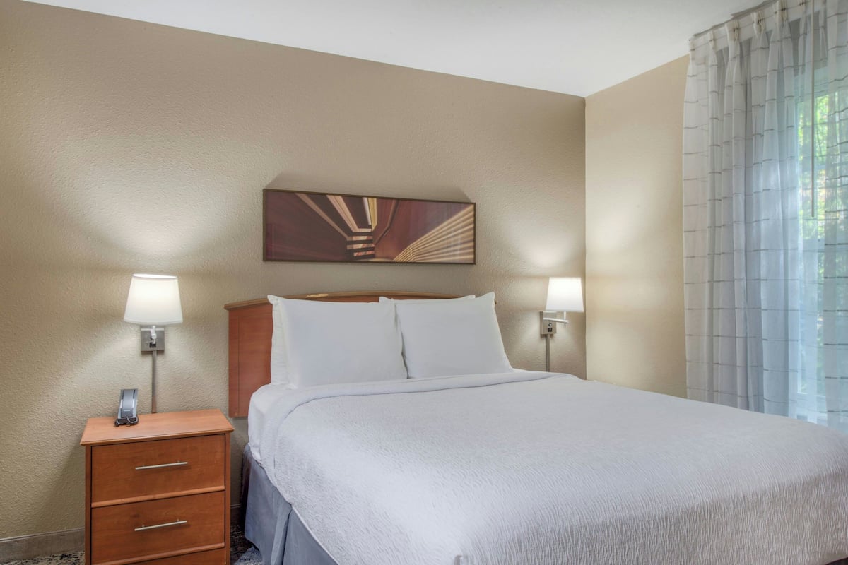 A hotel room with a bed, bedside lamps, a nightstand, a wooden headboard, and a painting on the wall.