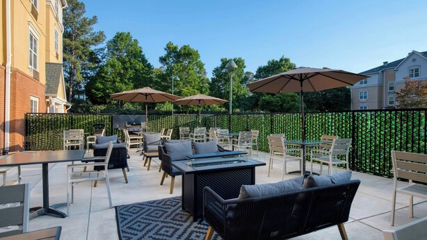 An outdoor patio with tables, chairs, and umbrellas, surrounded by a hedge and buildings. - TownePlace Suites by Marriott Raleigh Cary-Weston Parkway (Cary)