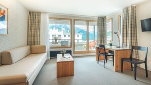 A modern hotel room with a sofa, a wooden desk with two chairs, a small coffee table, and large windows offering a mountain view.