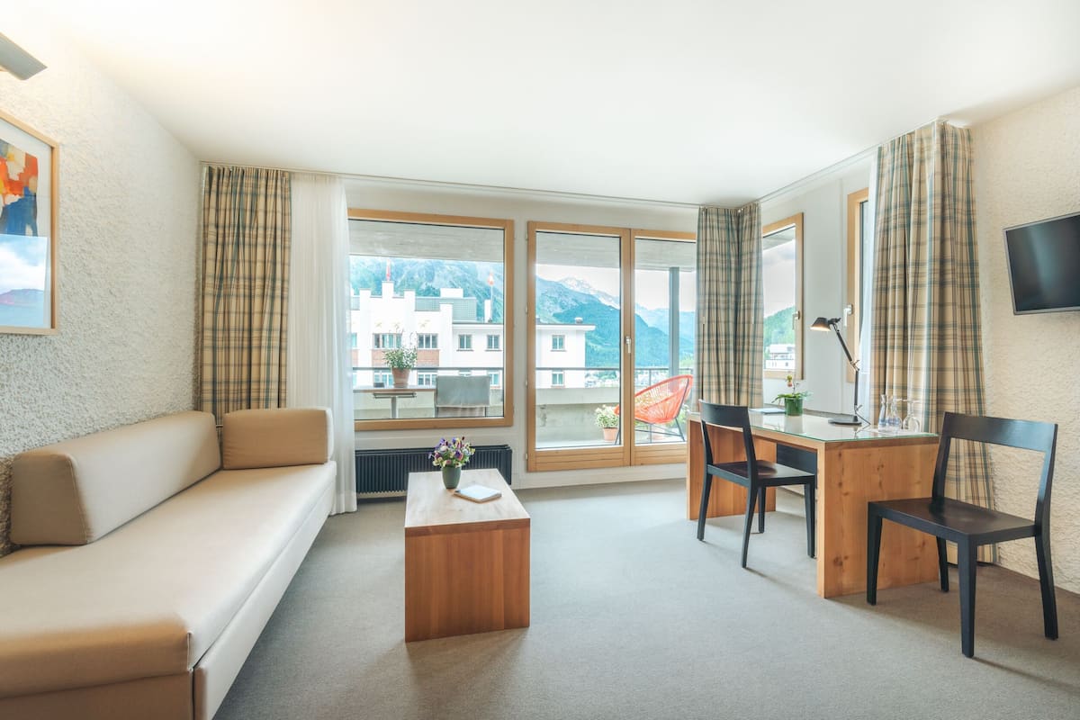Una habitación de hotel moderna con un sofá, un escritorio de madera con dos sillas, una mesita de centro y grandes ventanales con vistas a la montaña.