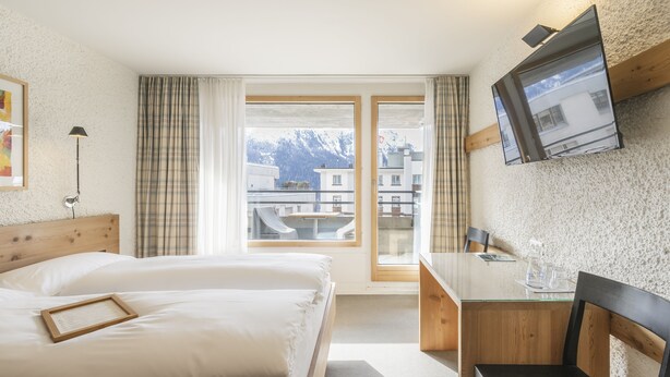 A hotel room with two beds, a desk, a chair, a TV, and a window with a view of buildings and mountains.
