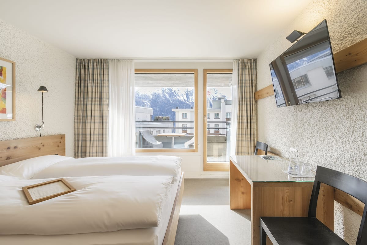 Habitación de hotel con dos camas, un escritorio, una silla, un televisor y una ventana con vistas a edificios y montañas.