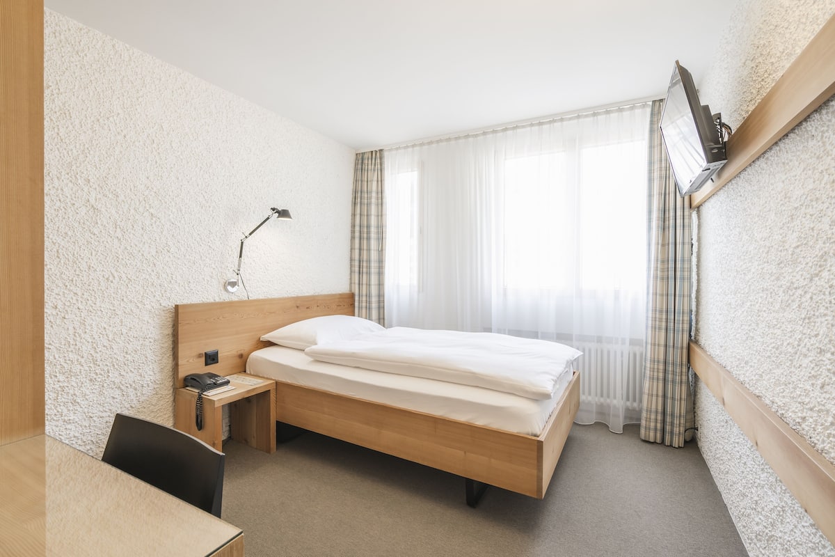 Habitación de hotel con una cama de madera, un escritorio con teléfono, una lámpara y una ventana con cortinas.