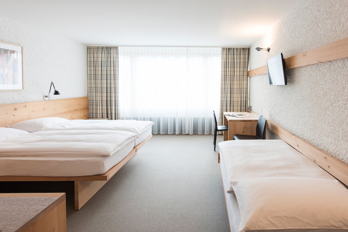 Habitación de hotel con dos camas, un escritorio, una silla, un televisor montado en la pared y una ventana con cortinas.
