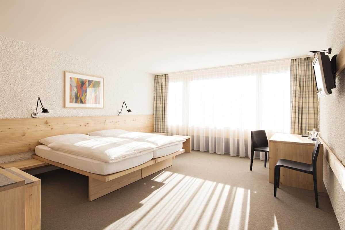 Habitación de hotel con cama de madera, un escritorio, una silla, televisor montado en la pared y una ventana con cortinas.