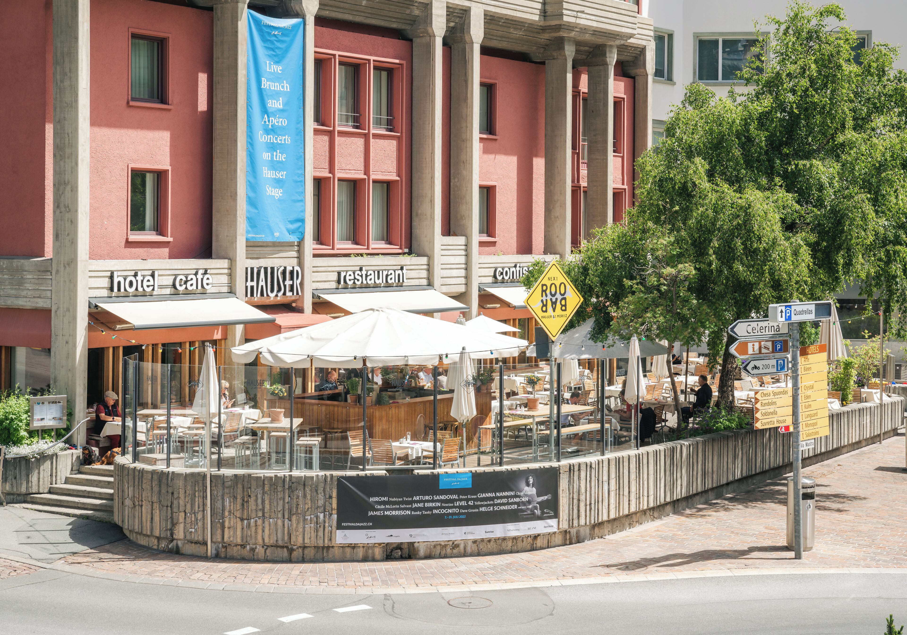 Un café al aire libre con mesas y sombrillas, un letrero que indica un café de hotel, y un edificio con columnas.
