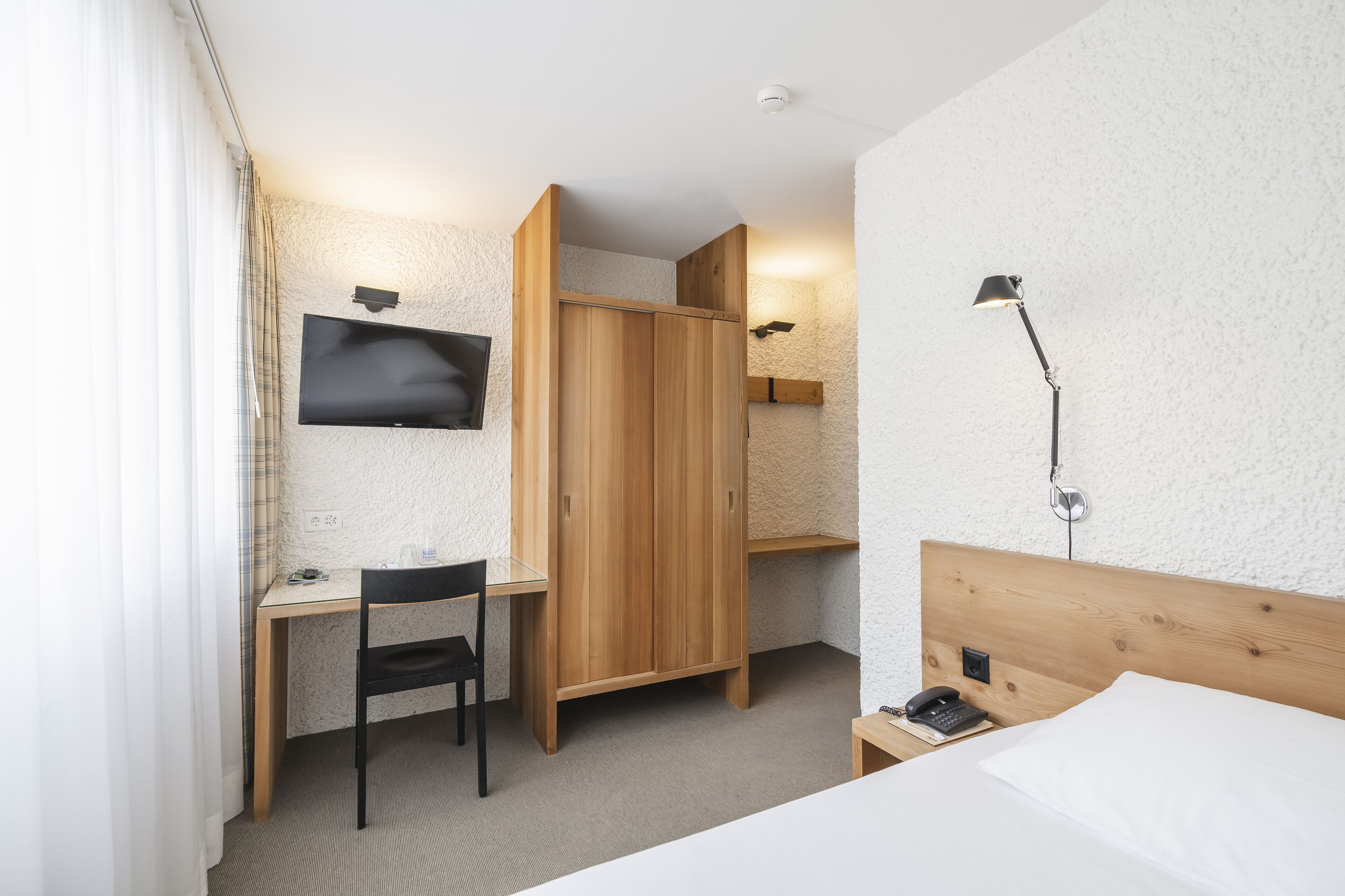A hotel room with a bed, a desk with a chair, a TV, a wardrobe, and a bedside table with a lamp.