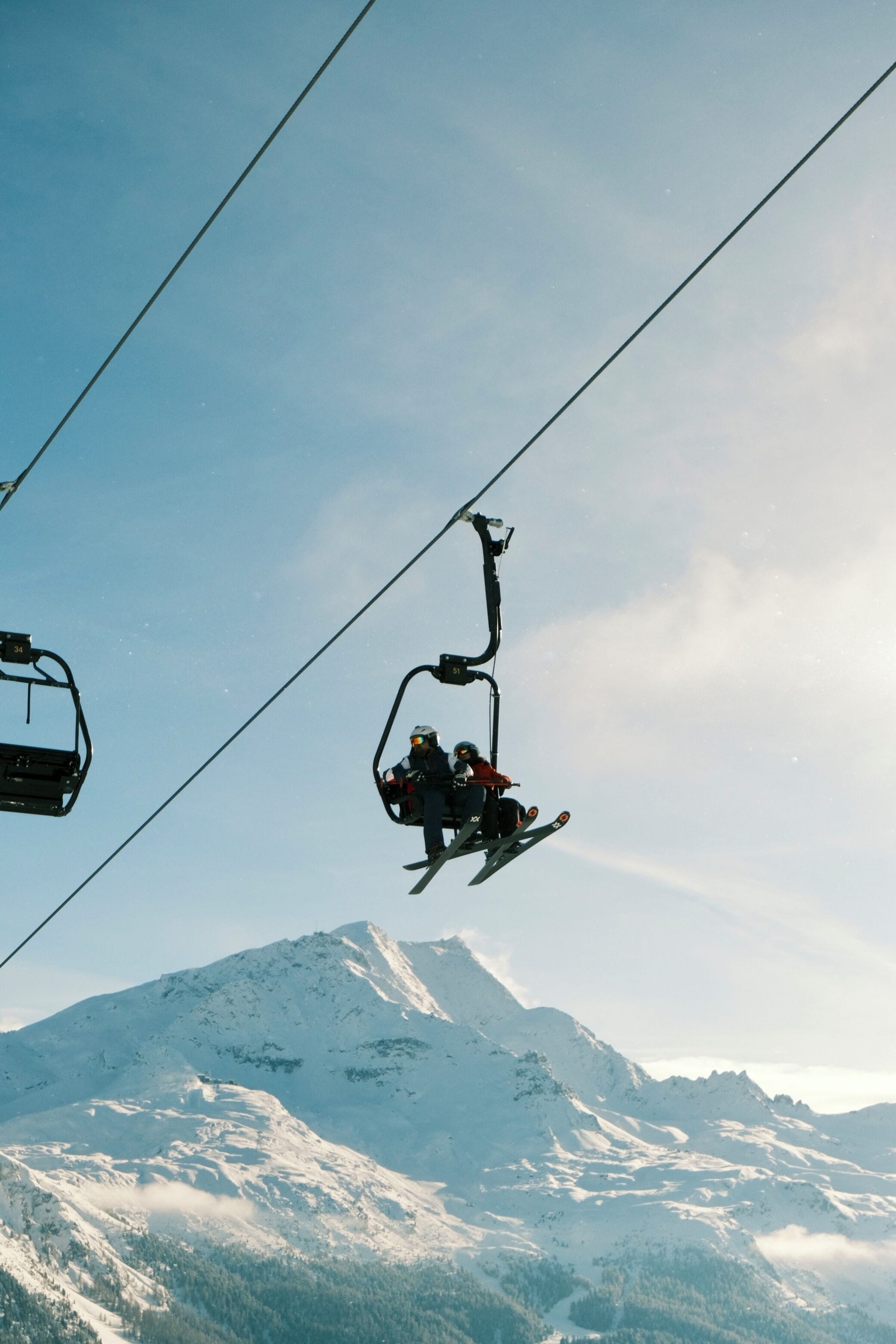 A ski lift with a person on a chairlift against a snowy mountain backdrop.