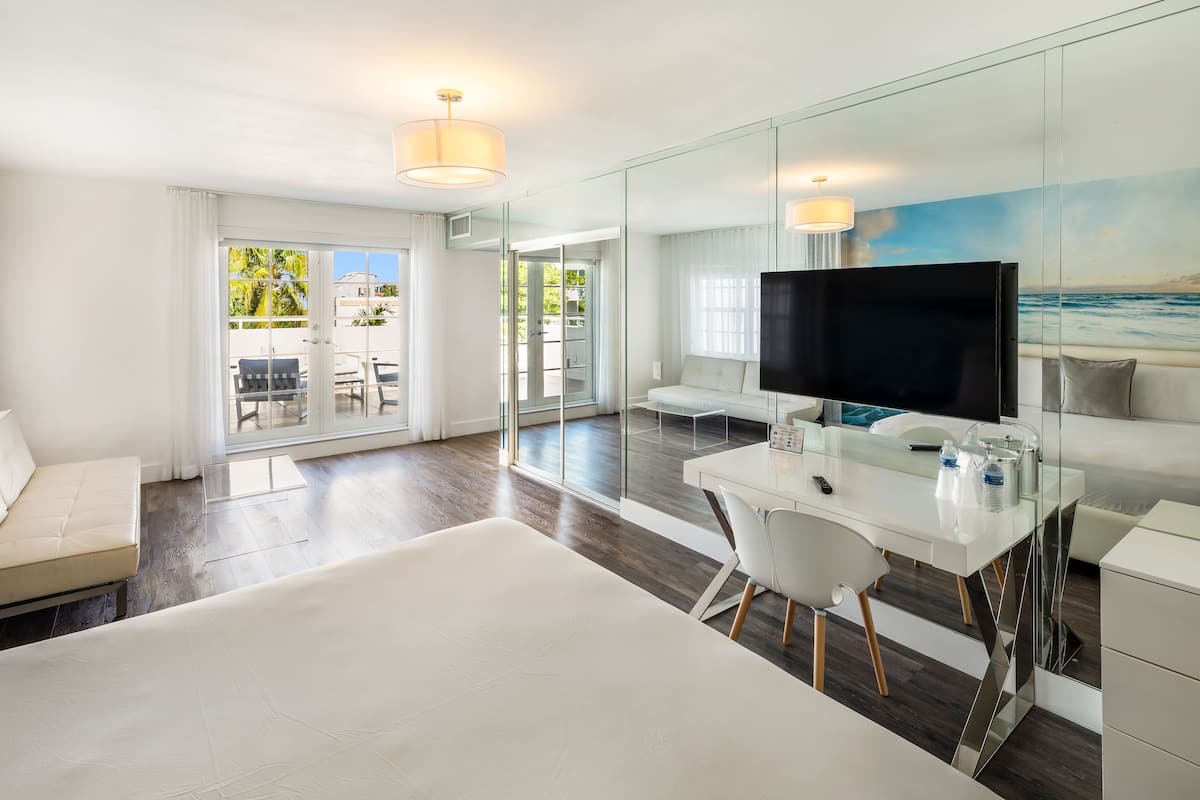 A modern hotel room with a large bed, a desk with a flat-screen TV, a glass partition, and a view of the outdoors.