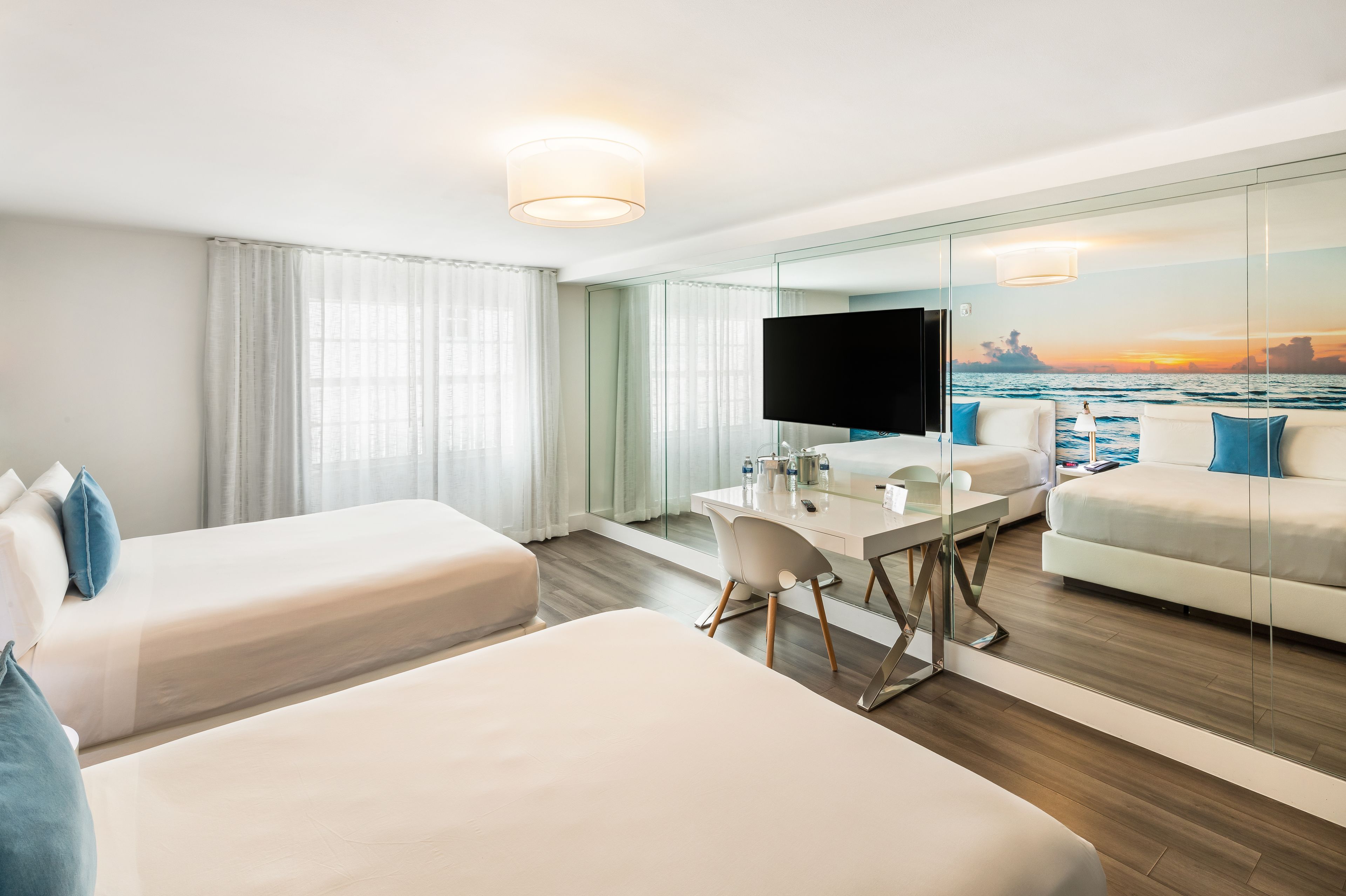 Habitación de hotel con cama, escritorio, silla, televisor y vista al océano a través de un muro de cristal.